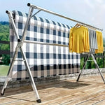 Foldable Clothes Rail displaying garments and a quilt on a sunny deck, showcasing the stainless steel X-frame design.