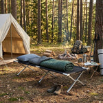 Heavy-Duty Portable Folding Camping Stretcher set up in a forest campsite with gear and a tent nearby.