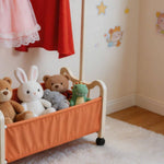 Colorful collection of stuffed animals in an orange storage bin, with a red dress hanging above on a steel clothing rack.