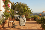 A grey wicker egg-shaped hanging chair, set against the backdrop of a South African garden terrace.