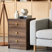 Modern 3-Drawer Nightstand with Gold Handles placed next to a grey sofa in a contemporary living room setting.