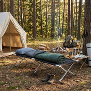 Heavy-Duty Portable Folding Camping Stretcher set up in a forest campsite with gear and a tent nearby.
