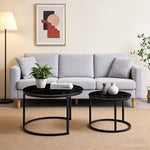 A styled living room scene featuring a light gray fabric sofa with two round black coffee tables in the foreground, along with a floor lamp, framed wall art, and a potted plant.