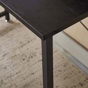 Close-up view of a sleek modern computer desk with a black top and sturdy metal legs on a textured floor.